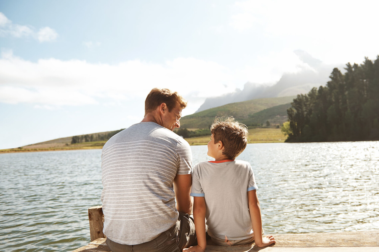 Rear view of a father and son bonding while they sit on a pier by a lake with copyspace