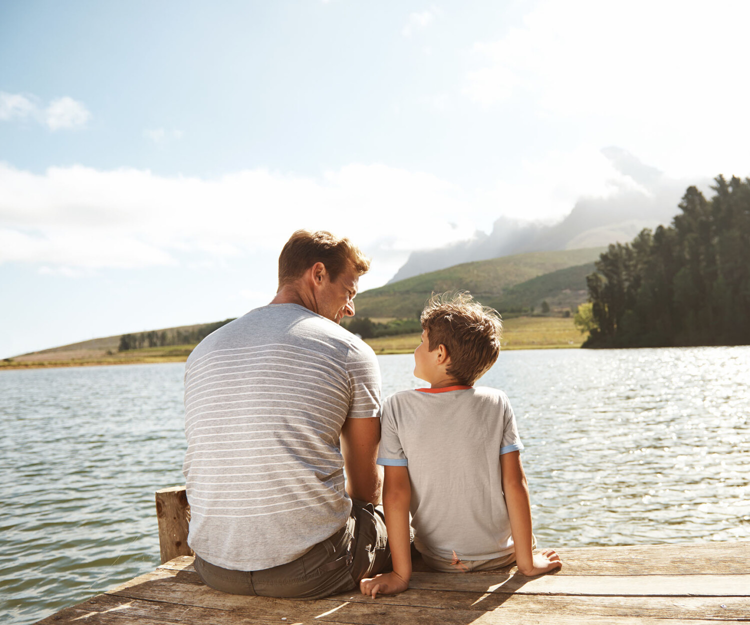 Rear view of a father and son bonding while they sit on a pier by a lake with copyspace
