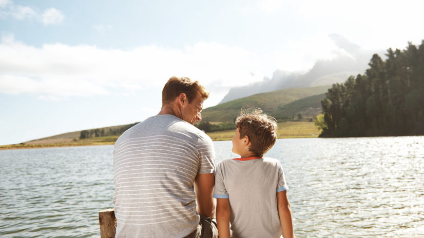 Rear view of a father and son bonding while they sit on a pier by a lake with copyspace